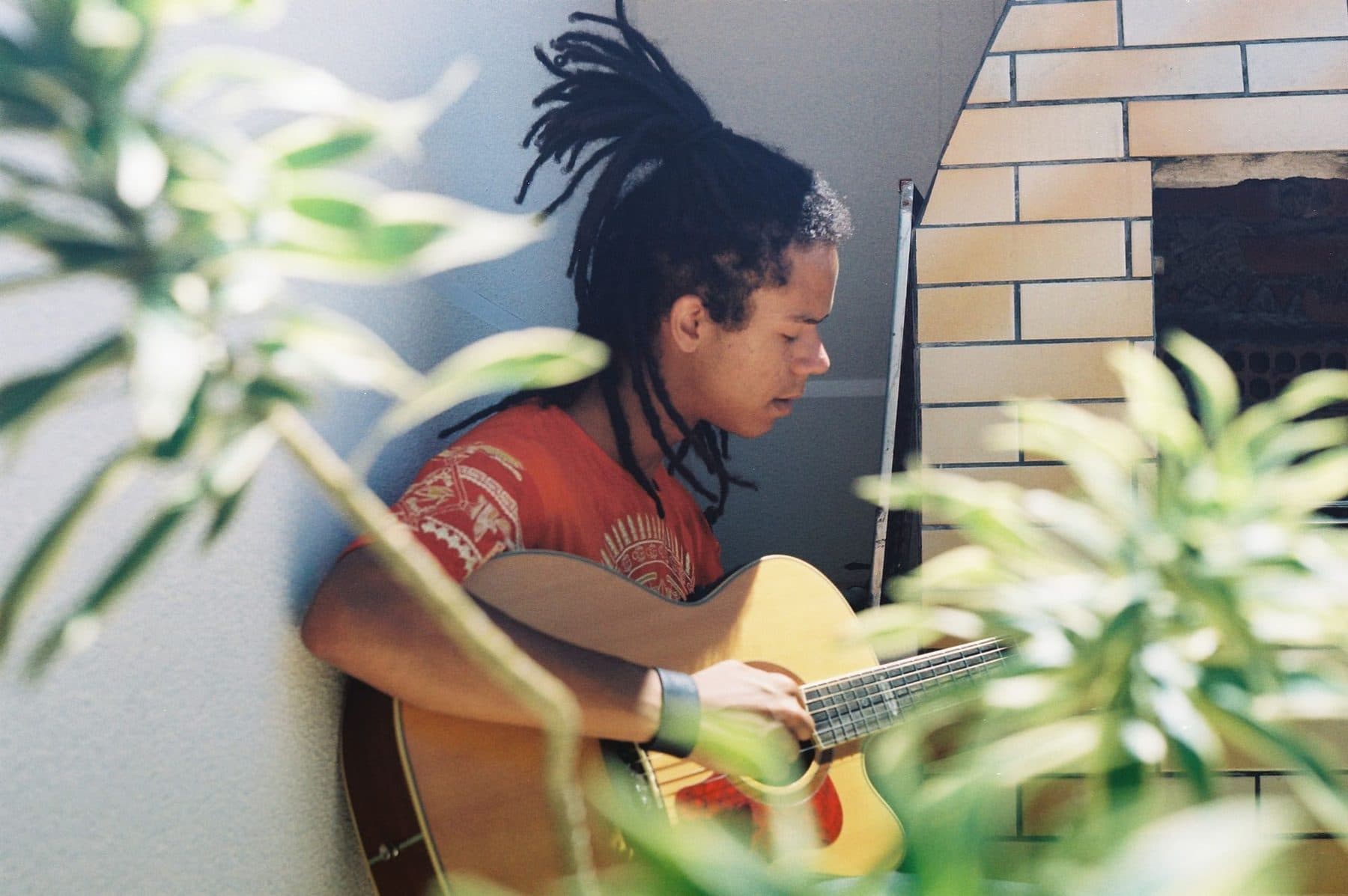 A teenager playing acoustic guitar outdoors, with green trees behind them.